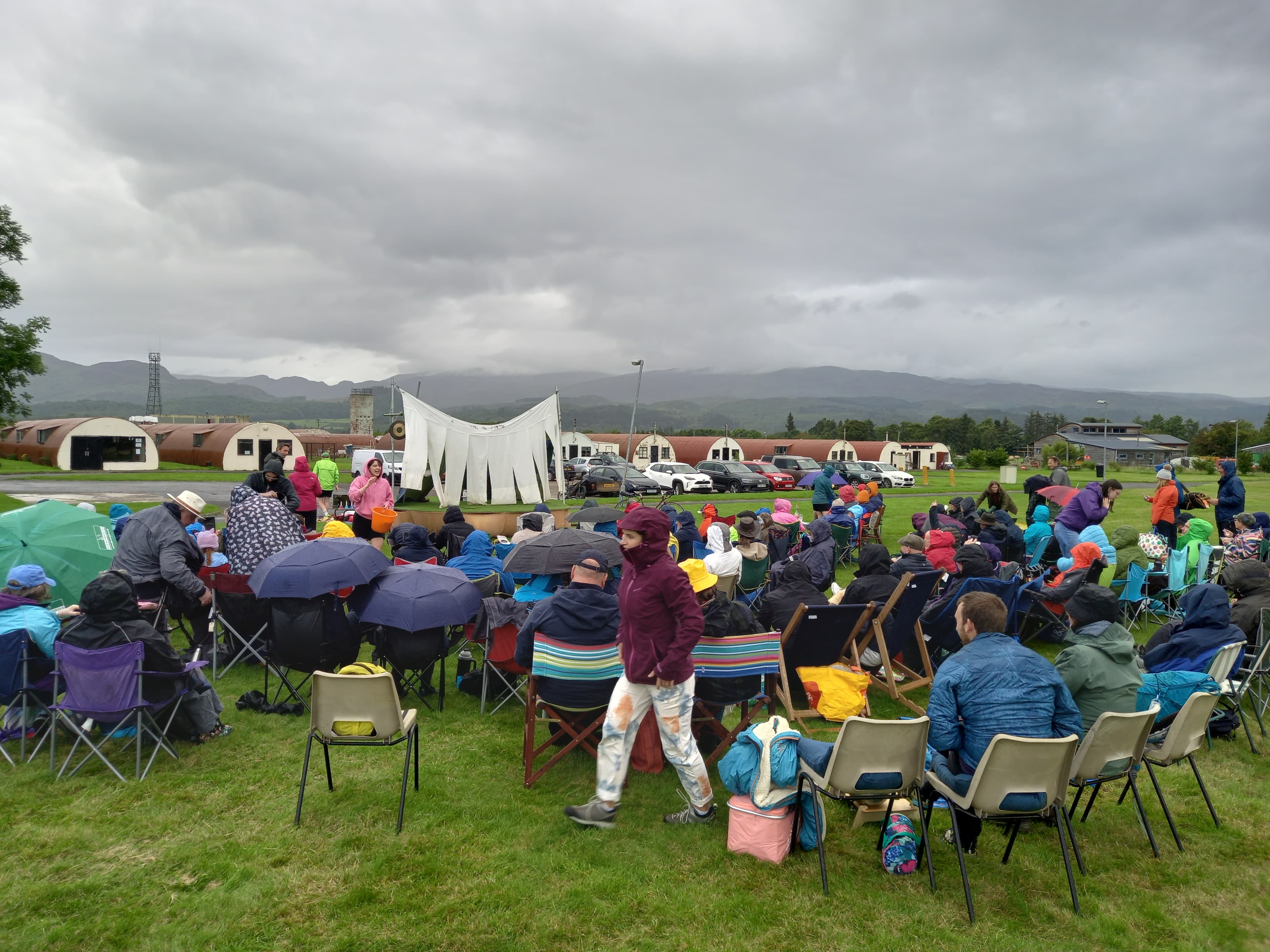 Audience seated on the grass watching outdoor theatre at Cultybraggan Camp with Nissen huts and hills behind